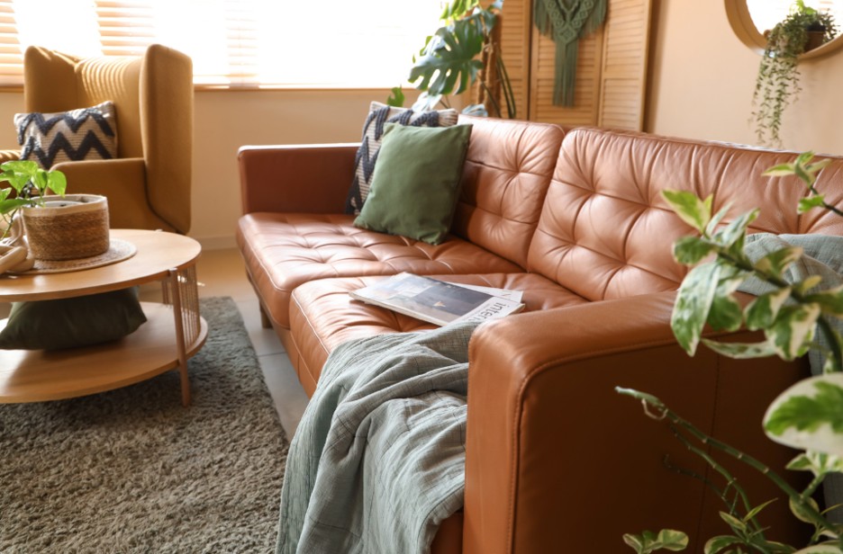 This is an image of an empty therapy room with a large brown leather sofa that is complimented with green accents, like a throw and pillow as well as plants. There is a coffee table as well and a large window