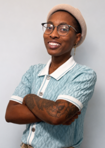 This is a profile picture of a therapist who is wearing a light blue collared shirt with a white collar. The person is also wearing a tan hat, glasses and has their arms crossed.
